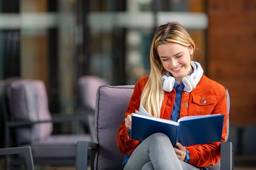 A cheerful student sits facing the camera, immersed in reading with an open book on her knees and headphones draped around her neck. Her joyful expression reflects her love for learning, as she eagerly absorbs knowledge from the pages before her. This image captures the essence of a student who finds happiness and fulfillment in the pursuit of education. Perfect for showcasing the enthusiasm and dedication that students bring to their studies in educational materials and promotional content.