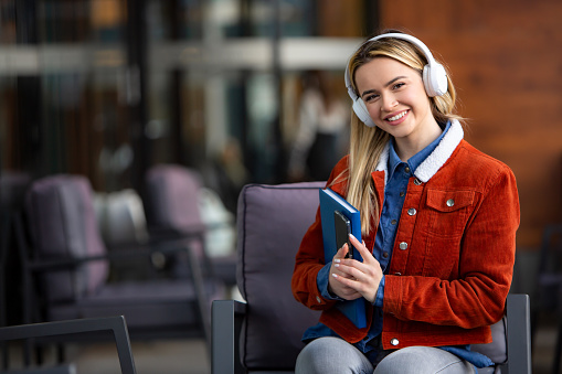 Front view, a beaming young blonde woman university adult student proudly holds closed books and a phone, adorned with headphones, signaling the end of her class with a big smile. Her radiant expression reflects the satisfaction of completing her studies, ready to unwind and enjoy her free time. This image captures the exuberance and sense of accomplishment that comes after a successful day of learning. Perfect for illustrating the joy and fulfillment of academic achievement in university promotions and educational materials.