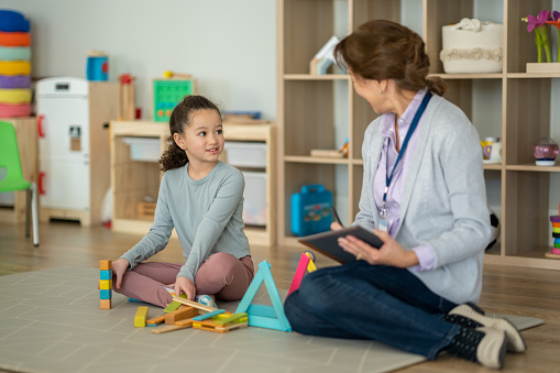 A female therapist of Asian decent, sits on the floor with a little girl during a play therapy session.  They have colourful blocks out in front of them and the therapist is taking notes electronically.