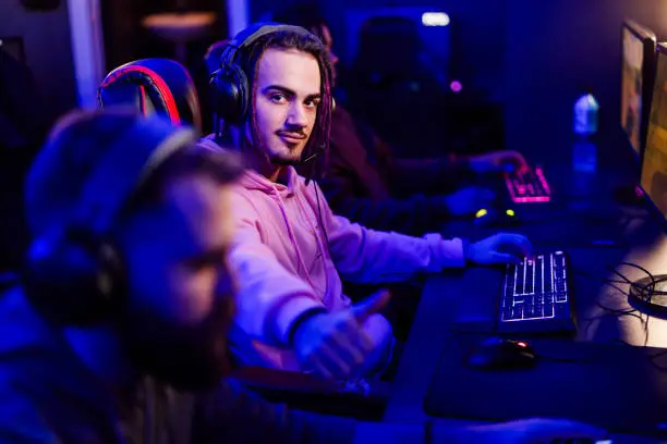 Portrait of a professional male with dreadlocks esport gamer wearing headphones and playing online video games while sitting in gaming club, internet cafe or cybersport lounge. Looking at the camera.
