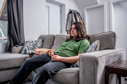 Indulging in a moment of leisure, a teenager immerses himself in the world of video games while relishing a slice of pizza on the comfort of his couch