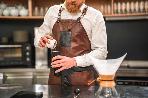 Close-up shot of a professional Caucasian male barista expertly weighing coffee beans on the counter using a digital scale in a coffee house cafeteria. Transferring coffee beans into a grinder.