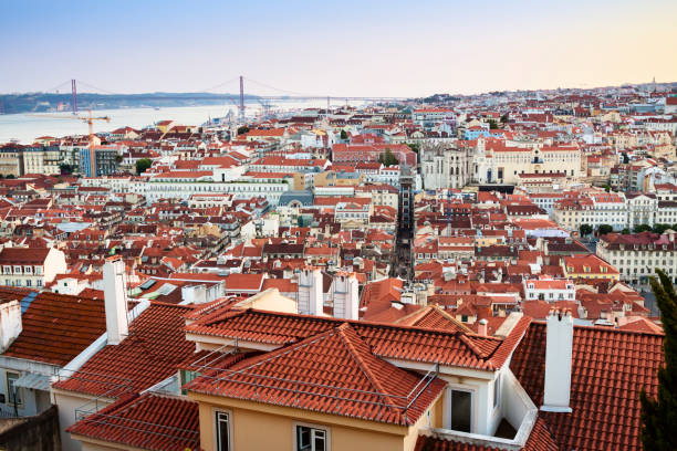 View over the downtown district of Lisbon at sunset with the Tagus river estuary in the background. The terracotta roof tiles dominate the foreground scene, the sky is clear and multicoloured as the sun goes down. The 25 April bridge can be seen on the horizon.
