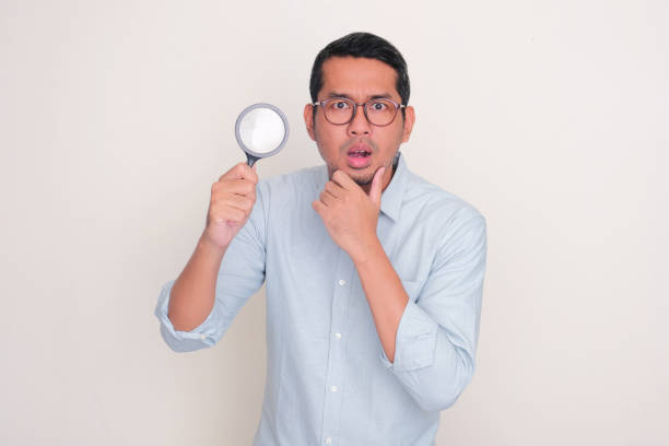 istock Adult man looking camera with suspicious expression while holding magnifying glass 2118483169