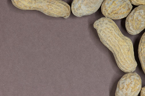 Unshelled peanuts scattered on brown cardstock background with copy space. Flat lay closeup food, legume crop nutrition and allergy, farming industry concept.
