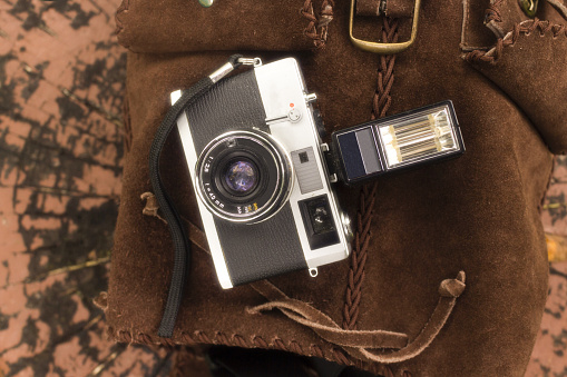 A classic rangefinder camera with a mounted flash sits atop a suede leather bag, surrounded by the warm, earthy tones of fallen autumn leaves. The scene evokes a sense of nostalgia and the art of traditional photography during a fall day.