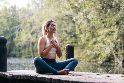Young woman in sportswear meditating and doing yoga in nature. She is barefoot on a wooden platform near a lake