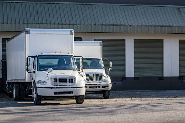 istock Day cab white middle duty rigs semi trucks with long box trailers standing in warehouse dock gates loading cargo for the next local deliveries 2118957611