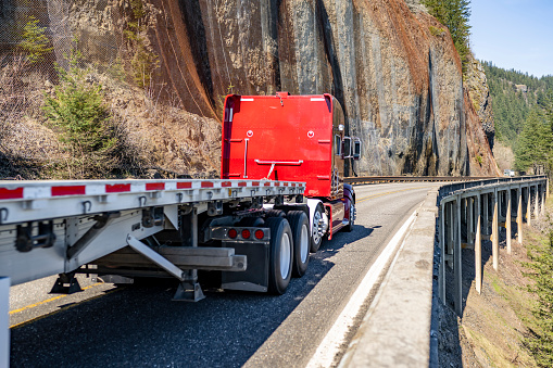 Powerful red big rig classic semi truck tractor transporting empty flat bed semi trailer running on the narrow mountain road with bridge on the slop