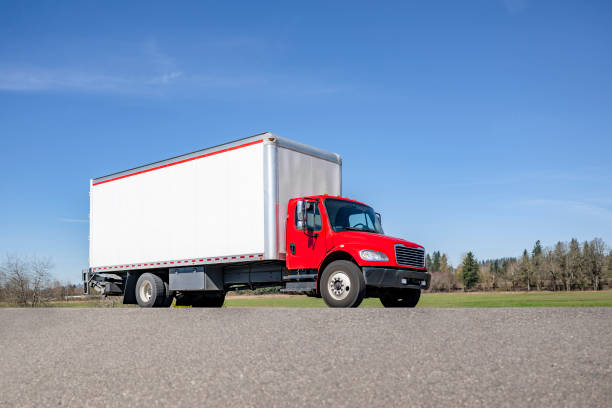 Middle duty industrial carrier red rig semi truck tractor with box trailer transporting cargo driving on the local road for delivery in sunny day