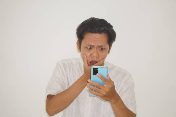 istock Young Asian man wearing a white shirt surprised while looking at smartphone isolated on white background 2119029391