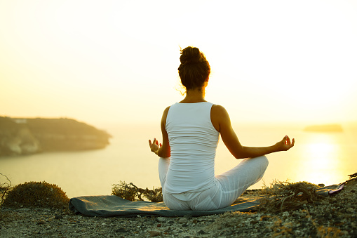 Rear view of a relaxed woman doing Yoga meditation exercises in Lotus position on a hill above the sea at sunrise. Copy space.