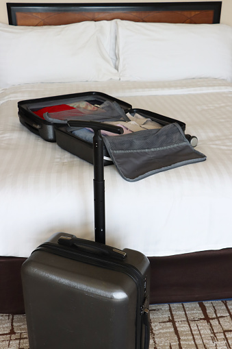 Stock photo showing close-up view of an open grey wheelie suitcase on a double bed mattress covered in fresh bedding, packed with neatly folded t-shirts and button-up shirts. A grey wheelie suitcase with a telescopic handle is at the foot of bed.