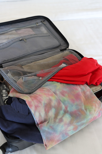 Stock photo showing close-up, elevated view of a grey wheelie suitcase on a double bed mattress covered in fresh bedding. The case has been packed with neatly folded t-shirts and button-up shirts.