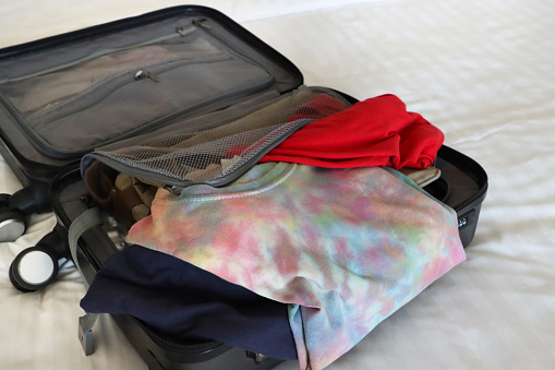 Stock photo showing close-up, elevated view of a grey wheelie suitcase on a double bed mattress covered in fresh bedding. The case has been packed with neatly folded t-shirts and button-up shirts.