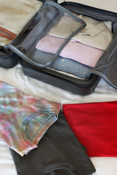 istock Close-up image of open grey wheelie suitcase on double bed, rows of packed folded collared shirts, luggage surrounded by t-shirts, neatly folded clothing, unzipped compartment mesh dividers, beige shoulder bag, elevated view, focus on foreground 2119188729