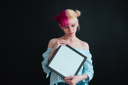 seriouse young  slim woman with pink hair holding empty frame for text or image in hands and looking on it. Waistup portrait on black background