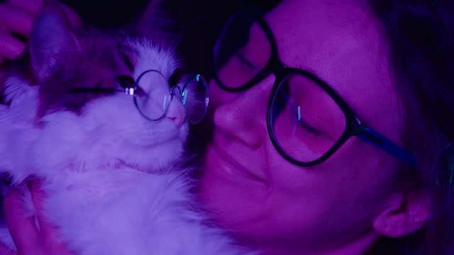 Bespectacled woman with cat hugging, cuddling, embracing. Blue-purple lamp illumination