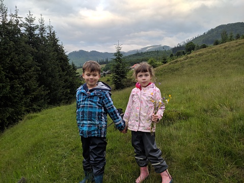 A girl and a boy are standing in a green meadow with a bouquet of wildflowers against the backdrop of green mountain peaks and tall fir trees.
