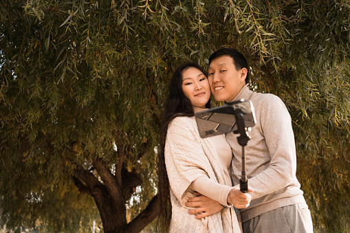 Happy hugging young asian couple in love taking selfie photo by phone under big green tree of weeping willow at summer day