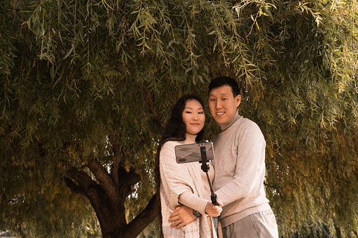 Happy hugging couple taking selfie photo by phone under big green tree walking in park at summer day