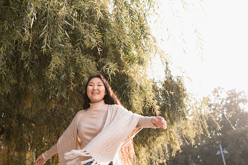 Happy young adult cheerful asian woman spinning around and dancing in a park having fun in sun rises