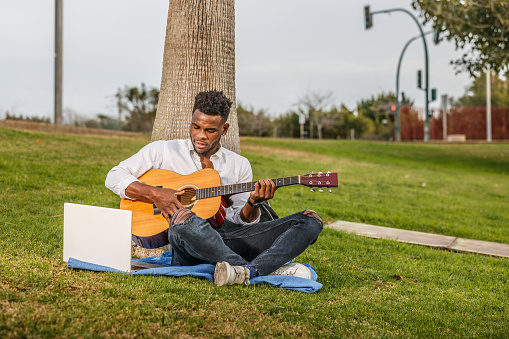 A man in casual clothes is sitting on the green grass and playing a guitar.