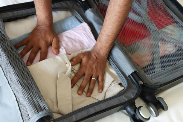 Full frame image of unrecognisable man packing grey wheelie suitcase on double bed covered by white duvet,  leaning over to pack folded collared shirts, neatly folded clothing, compartment mesh divider, buckled adjustable tension strap, elevated view
