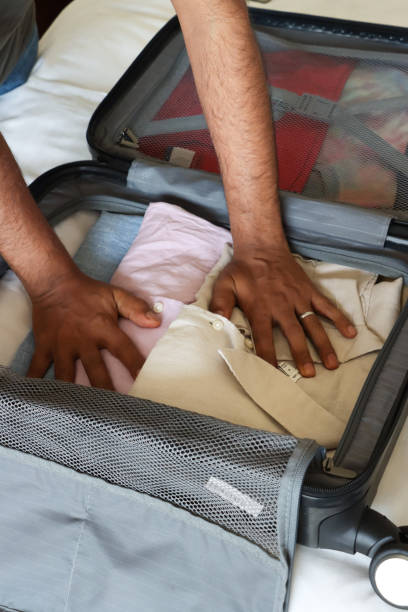 istock Closed image of unrecognisable man packing grey wheelie suitcase on double bed covered by white duvet,  leaning over to pack folded collared shirts, neatly folded clothing, compartment mesh divider, 2119303556