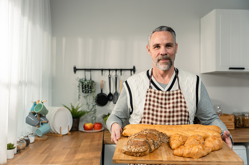 Portrait of Caucasian senior man hold tray of bread, looking at camera. Attractive elderly mature grandfather wear apron, spend time cook food breakfast for health care in house. Diet and Healthy food