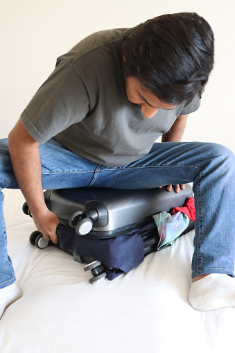 Stock photo showing close-up view of a grey wheelie suitcase on a double bed mattress covered in fresh bedding. The case has been packed with t-shirts.