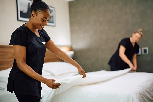 Hotel maids making bed in guest room. Focus is on African American maid.
