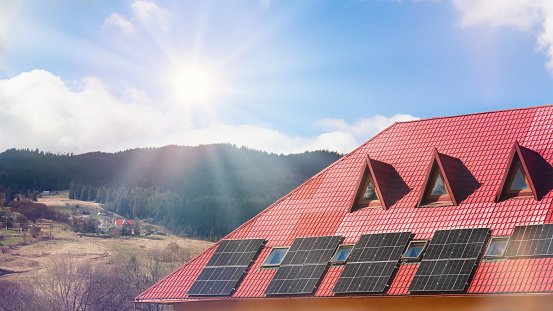 Solar panels installed on red-roofed house with scenic mountain backdrop and clear blue sky with shining sun.