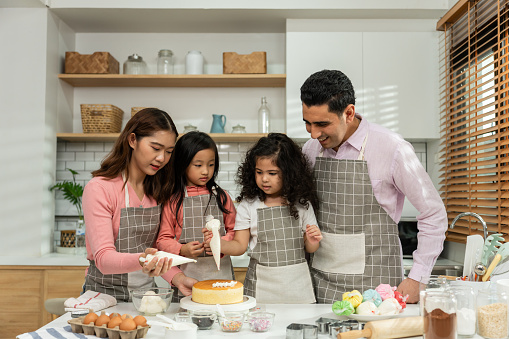 Asian attractive couple making cake with daughters in kitchen at house. Happy Family, parents and young kid girls having fun spending time together using ingredient bake bakery. Activity relationship.