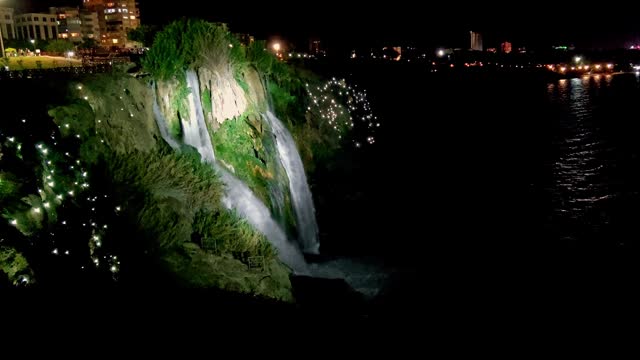 Nght Waterfall with Beautiful Lighting Flowing into Sea from Great Height of Steep Shore, against Background of Night City Lights., Concept of Merging City with Nature