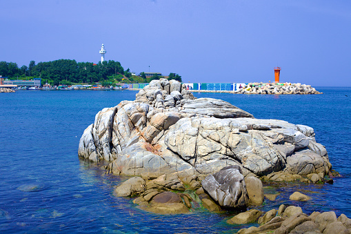 Goseong County, South Korea - July 31, 2019: Overlooking the serene waters of Daejin Port, a large rock emerges from the East Sea, with a small red lighthouse marking the breakwater's end and the majestic Daejin Lighthouse standing guard on the hill above.