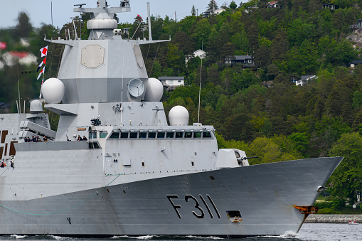 One crew member is raising two maritime flags, while the other watches or assists, on the bridge of USS Canberra (LCS 30), an Independence Class combat ship of the US Navy which is docked at Garden Island in Sydney Harbour.  She was in port for her commissioning ceremony on 22 July 2023.  As part of the ceremony, her captain was presented with a kangaroo insignia attached to all ships of the Royal Australian Navy, but decorated in the colours and design of the Stars and Stripes.  That insignia has been attached to the ship and visible on the right.  The name of the ship and serial number is visible on the orange life ring.  A large, padded chair is visible on the bridge.  This image was taken on a cloudy afternoon on 29 July 2023.