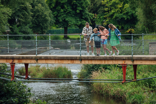 Wide shot of a family of four standing together on a bridge. The family consists of a mother and father with their son and daughter. The bridge is across a river located in Morpeth in the North East of England. They are looking down at the water. 

Videos are available similar to this scenario.