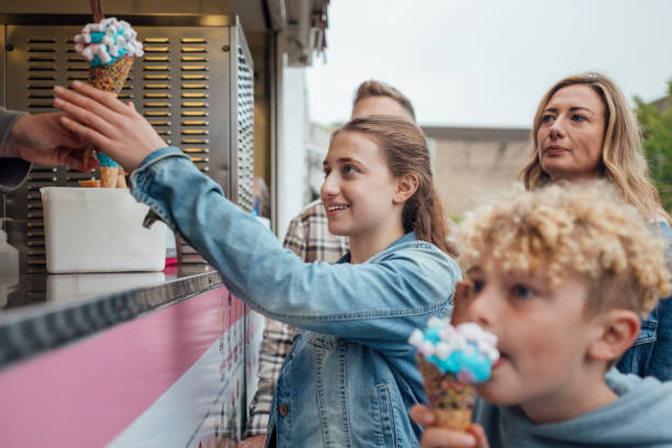Medium shot of a young boy eating an ice cream. His family is watching him eat the ice cream while smiling. The ice cream is blue and pink with toppings on it. His sister is receiving an ice cream from the ice cream stand.\n\nVideos are available similar to this scenario.