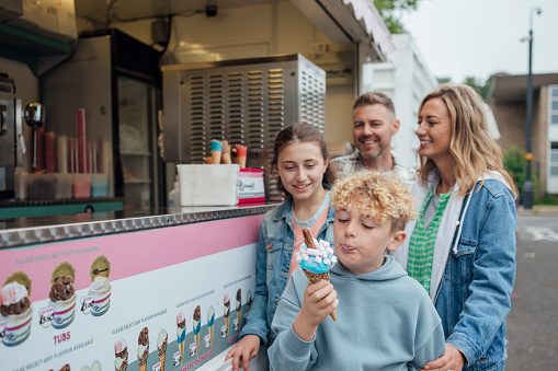 Medium shot of a young boy eating an ice cream. His family is watching him eat the ice cream while smiling. The ice cream is blue and pink with toppings on it. The family are waiting behind the boy for their ice cream.

Videos are available similar to this scenario.