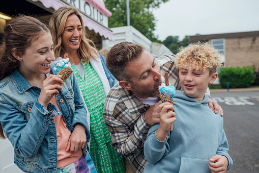 Medium shot of a young boy eating ice cream with his sister. His family is watching him eat the ice cream while smiling. The ice cream is blue and pink with toppings on it. His father is taking a lick of his son's ice cream.\n\nVideos are available similar to this scenario.