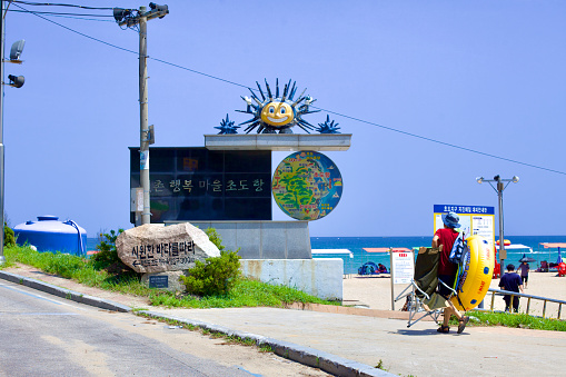 Goseong County, South Korea - July 31, 2019: At the north end of Hwajinpo Beach, a visitor prepares for a day by the sea, passing a granite sign with a sun statue, directing towards Chodo Port and beach.