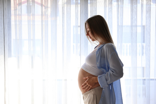 Young pregnant woman standing by the window and touching her belly in soft natural light. Close up, copy space, background.