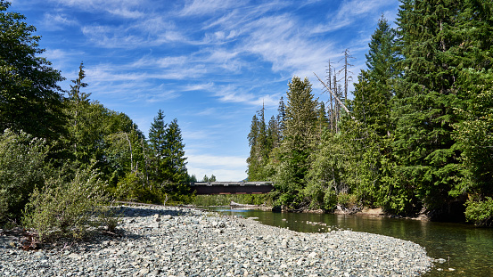 Looking down a river to a remote logging road bridge with wispy clouds in the blue sky.