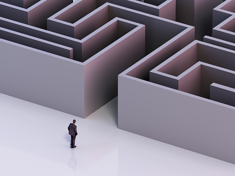 A solitary businessman in a suit stands at the entrance of a large, intricate maze, pondering the path he should take through the towering walls that lie ahead, symbolizing strategic decision-making in a challenging environment.