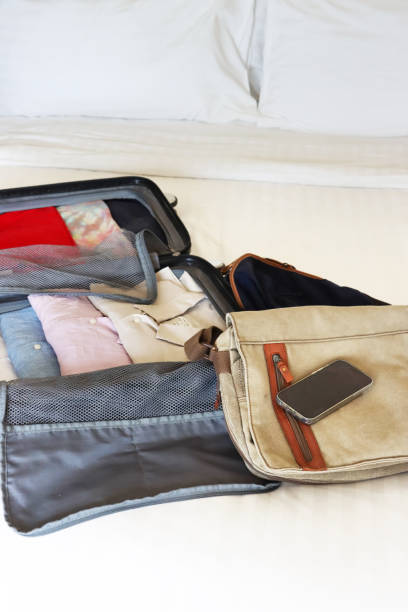istock Image of open grey wheelie suitcase on double bed covered by white duvet, rows of packed folded collared shirts and t-shirts, neatly folded clothing, unzipped compartment mesh dividers, blue rucksack and beige shoulder bag, Smartphone, focus on foreground 2119651675