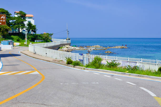 Goseong County, South Korea - July 31, 2019: The winding seaside road at Daejin Port, bordered by a white fence separating it from the East Sea, offers a picturesque journey with views of offshore rocks.