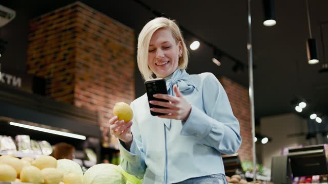 Blonde woman standing by showcase with fresh fruits while holding smartphone and lemon in hands. Smiling female customer nodding approvingly as sign of satisfaction with quality of groceries.