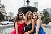 Three beautiful happy elegant girl friends with a smile in fashion dresses sitting near the fountain in the city