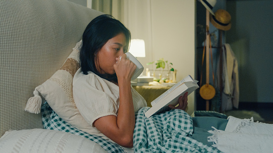 Young Asian girl reading book and drinking milk sitting on the bed in home at night. Insomnia, Cybersickness, Nomophobia, sleep disorder concept.